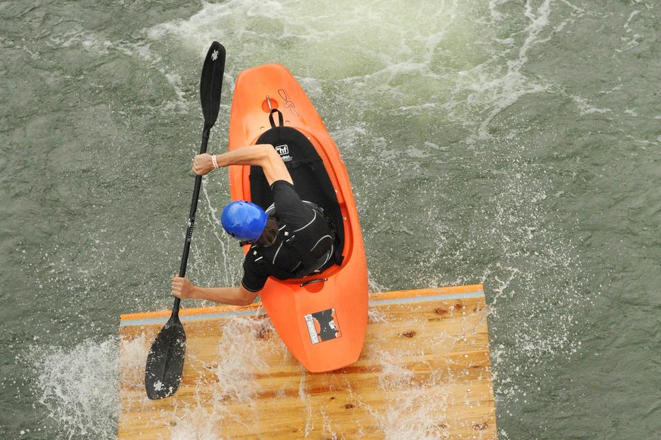 Paddler in orangenem Kajak steuert über eine Holzrampe ins Wasser, spritzendes Wasser um ihn herum.